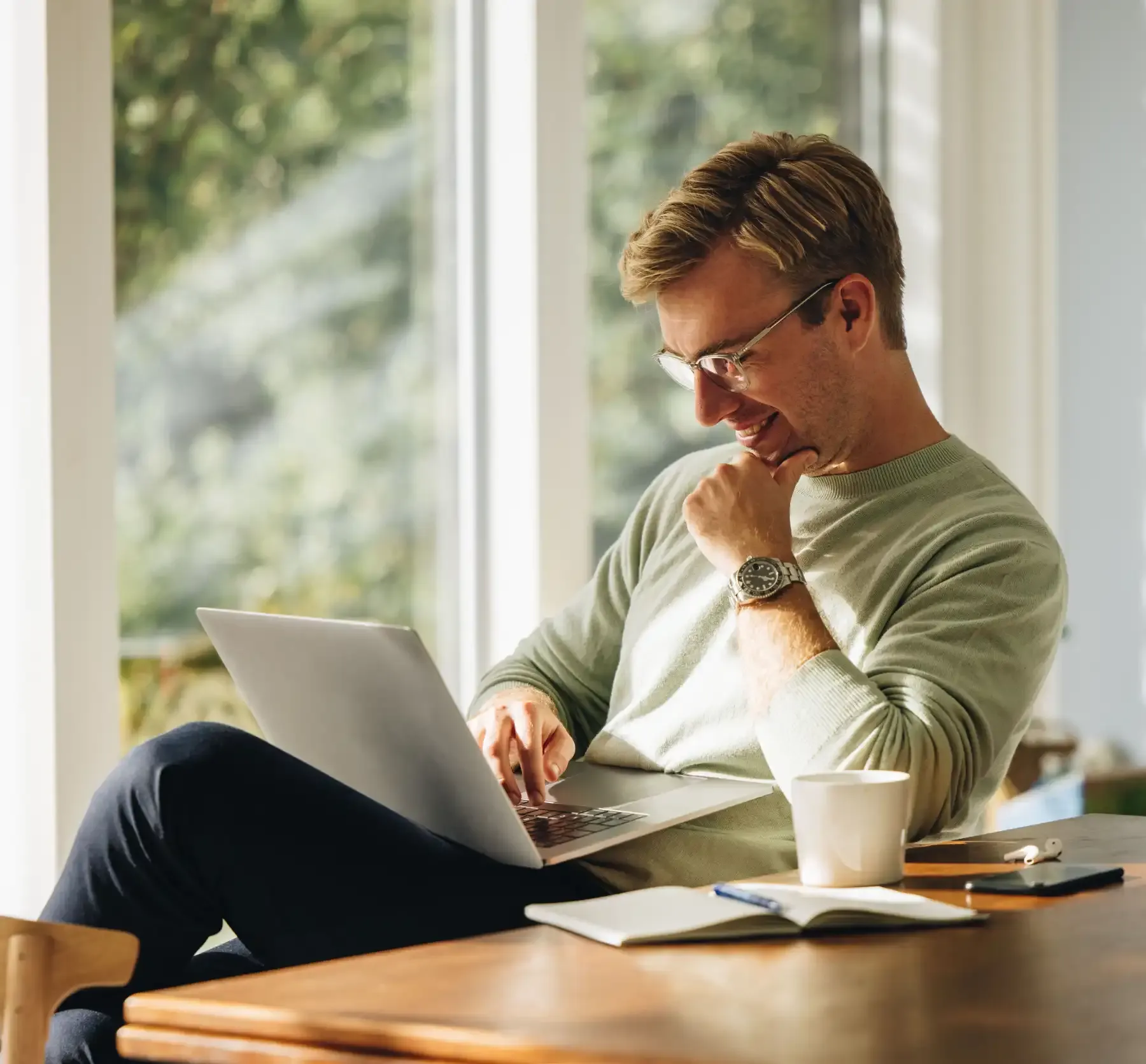 En mann sitter og ser på en laptop, smilende, lys inn gjennom vinduet i bakgrunnen